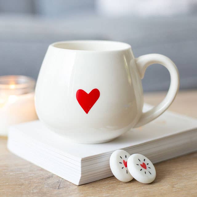 A white mug with a red heart on the front, resting on a stack of books.