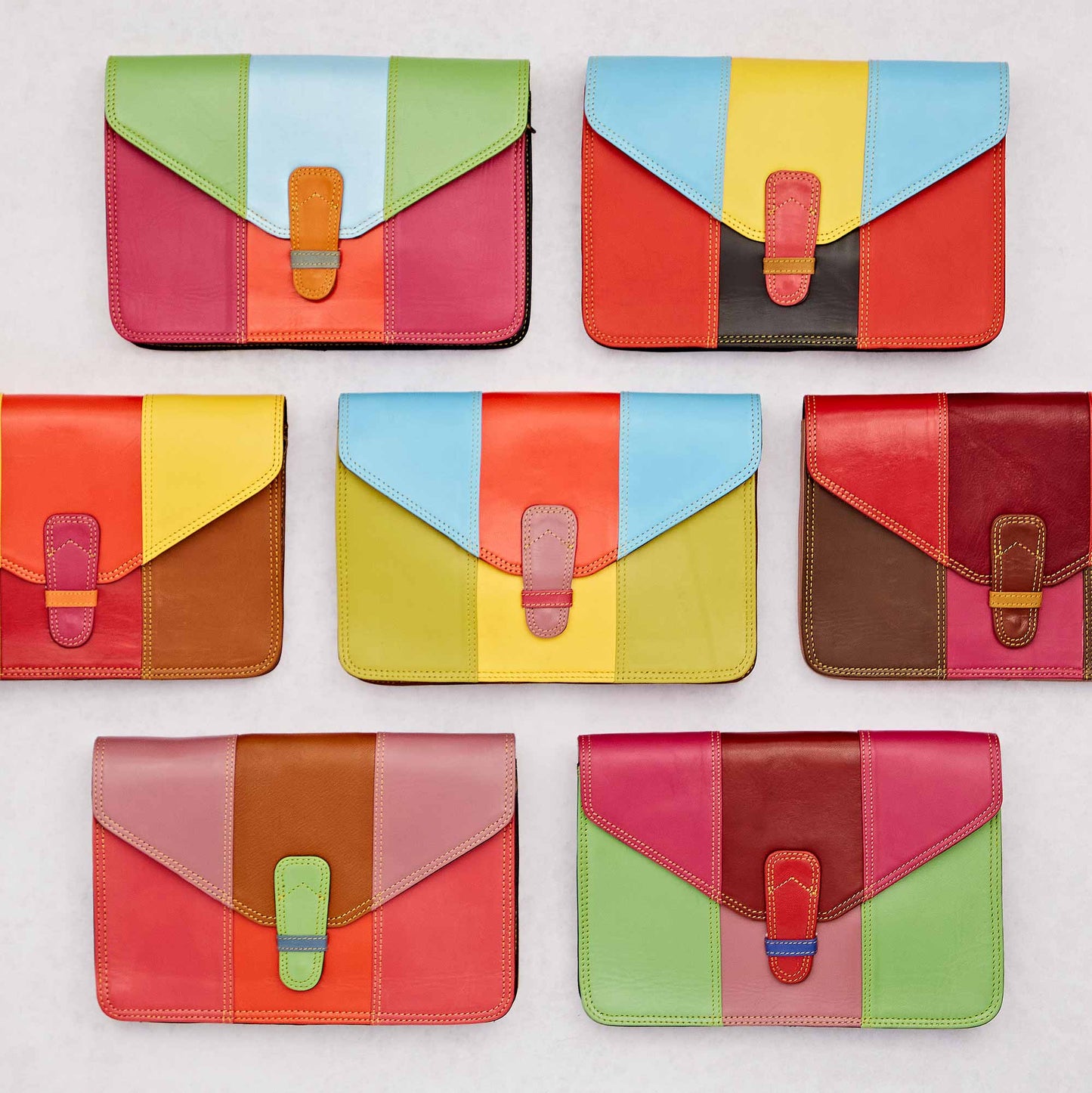 A collection of vibrant, multicolored recycled leather clutch bags arranged on a light background. Each bag features a unique patchwork design with bold colour blocking and a secure strap closure.