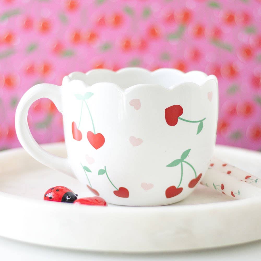 A close-up of a white mug with a scalloped rim. The mug is decorated with small red cherries with green stems and pink hearts.