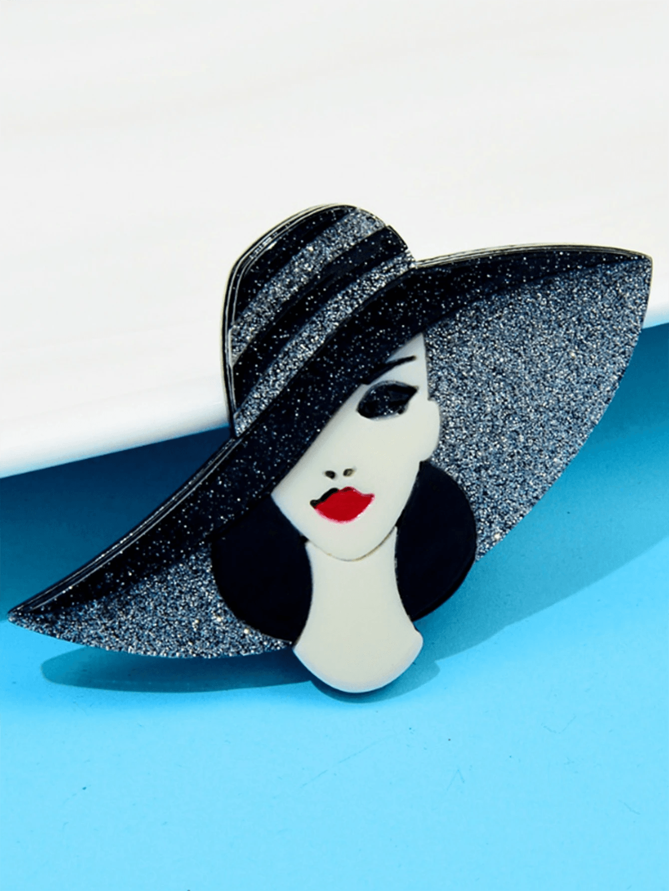 Handmade acrylic brooch with a stylish lady with a glittery black wide-brim hat, red lips, and an elegant design on a light blue background.