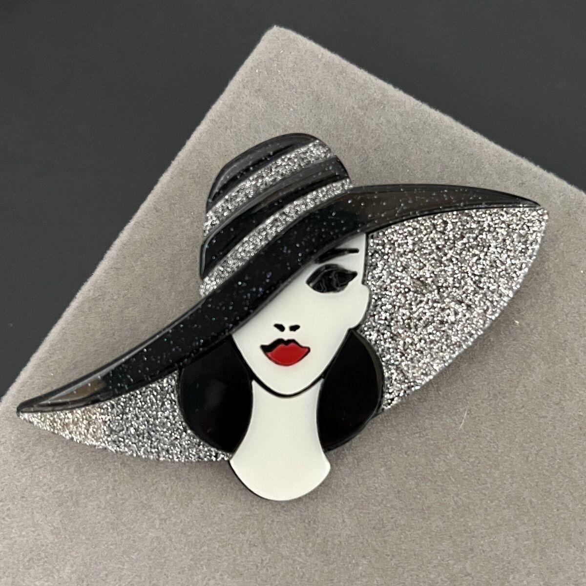 Handmade acrylic brooch featuring a stylish lady with a glittery black wide-brim hat, red lips, and an elegant design on a grey background.