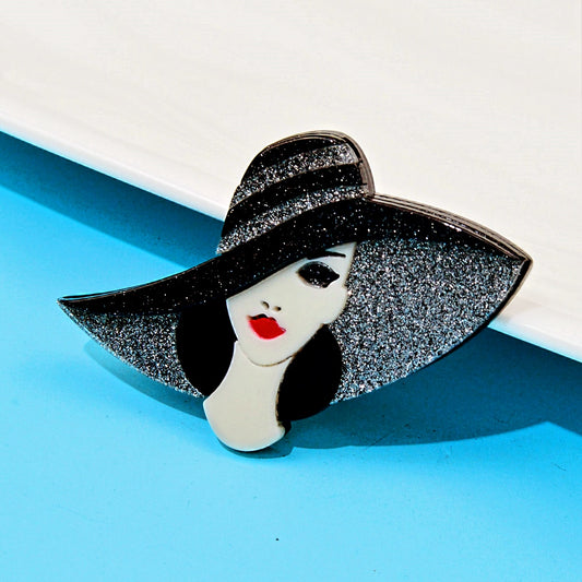 Handmade acrylic brooch featuring a stylish lady with a glittery black wide-brim hat, red lips, and an elegant design on a light blue background.