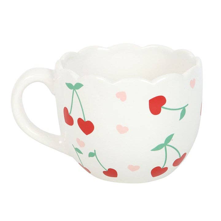 A close-up of a white mug with a scalloped rim. The mug is decorated with small red cherries with green stems and pink hearts. White background.