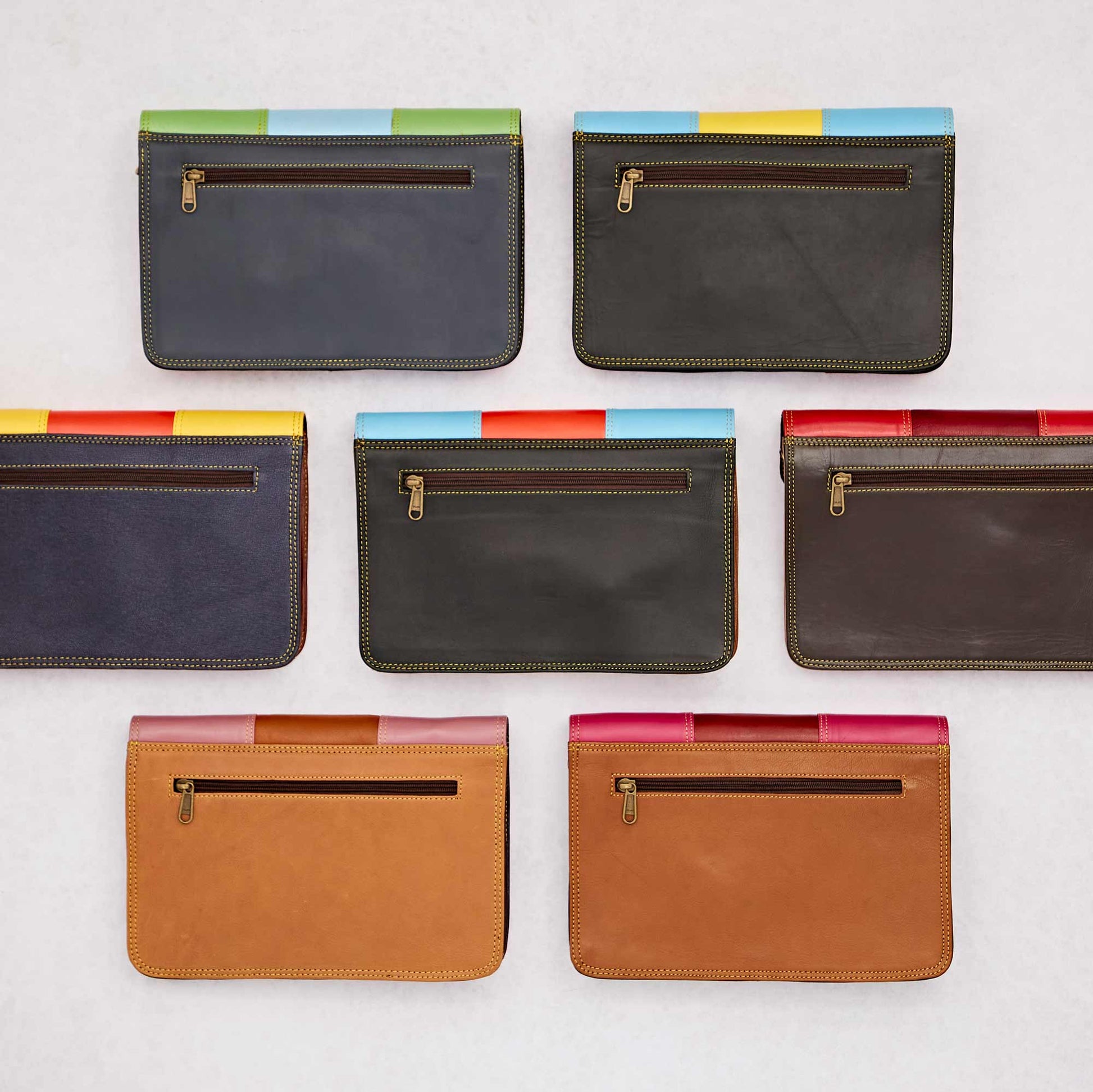  A collection of recycled leather clutch bags displayed with their backs facing up, showcasing zippered pockets and colourful trim details. The bags feature a mix of dark and neutral tones with vibrant accents along the top edge.