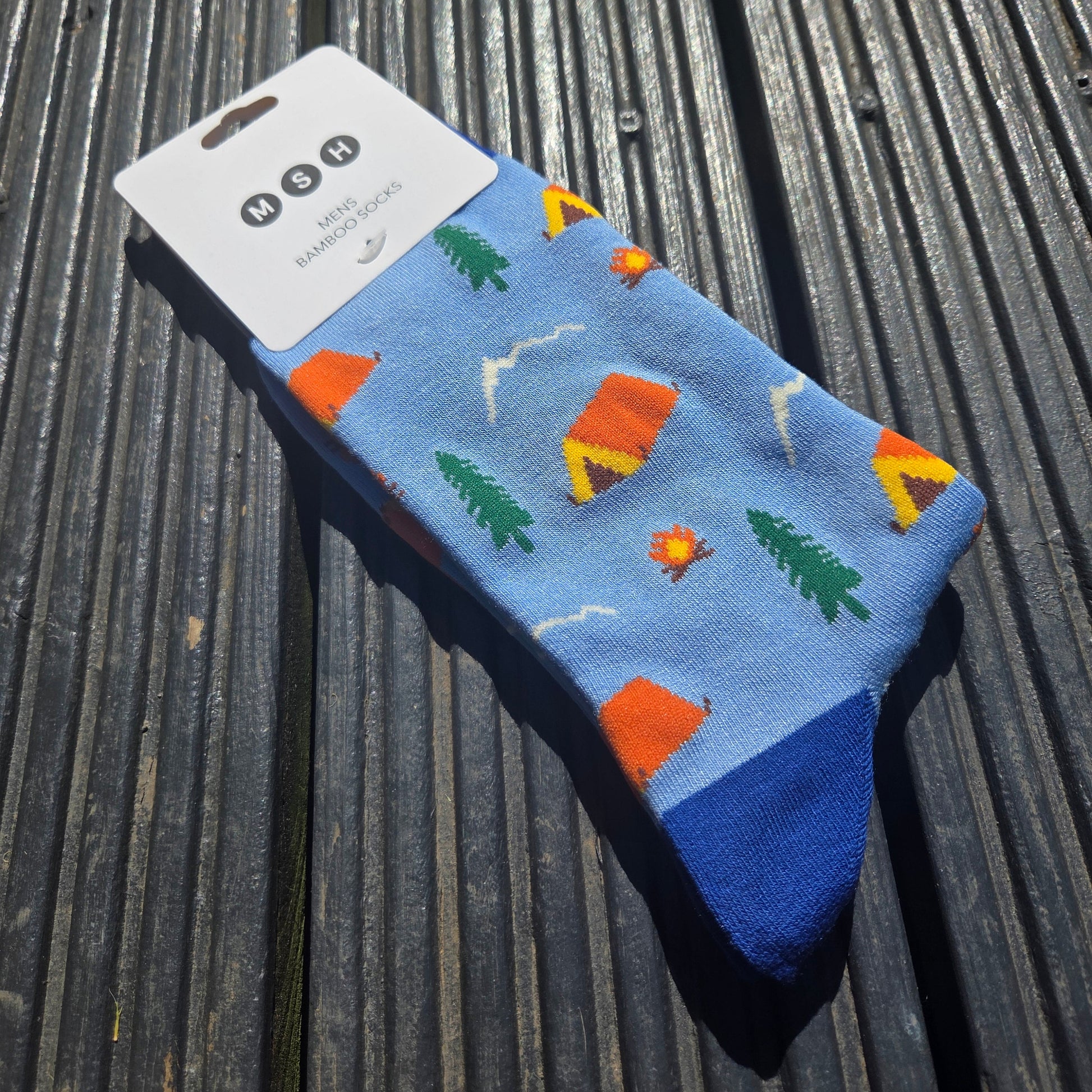 A pair of light blue bamboo socks featuring a campsite scene with tents and trees on a dark grey wooden background