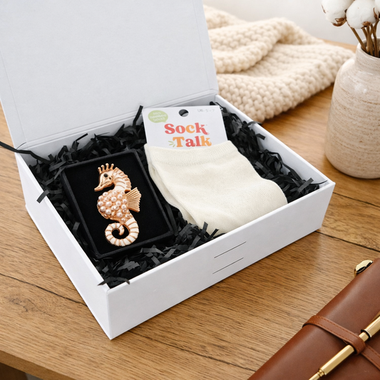 Open white gift box with rose‑gold seahorse brooch and Sock Talk bamboo socks on wooden table.