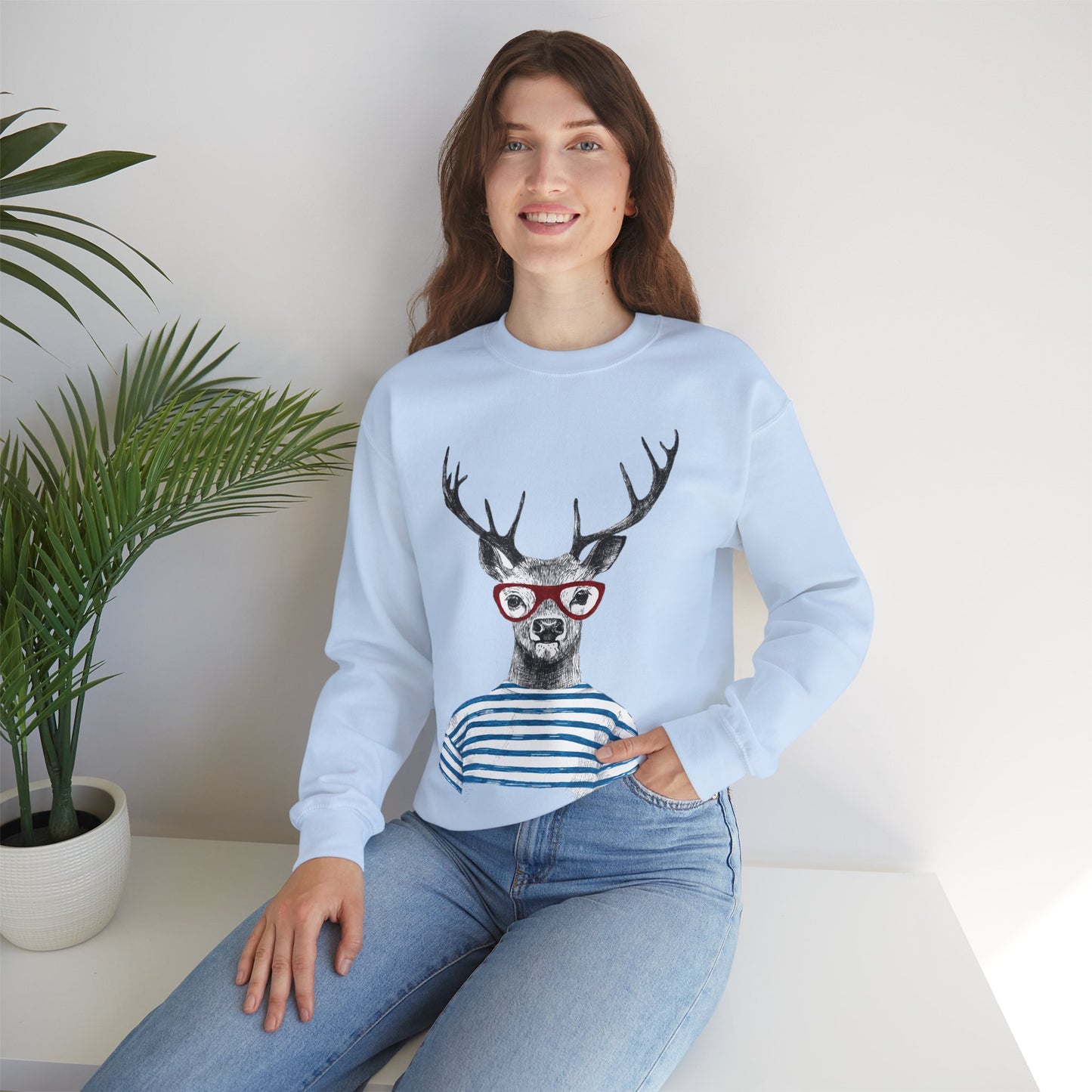 Woman wearing a Wild & Free Reindeer unisex sweatshirt in Light Blue with quirky stag print, sitting on a white surface with a plant in the background.