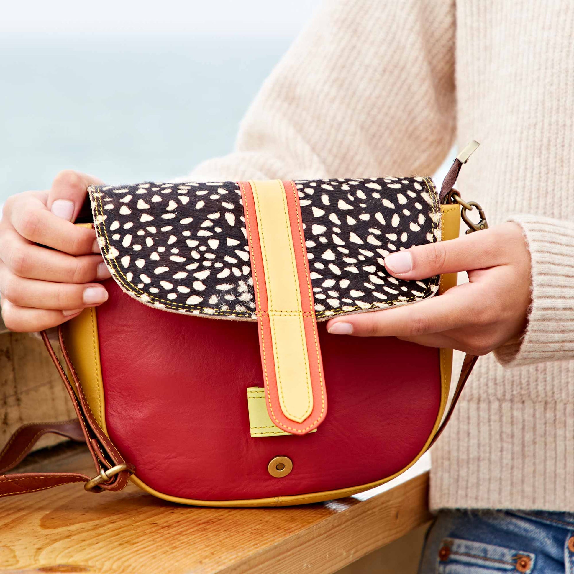 A person wearing a beige knit sweater and jeans holds a red handbag with yellow trim and a black‑and‑white animal‑print flap, standing near a wooden surface with a blurred seaside background.